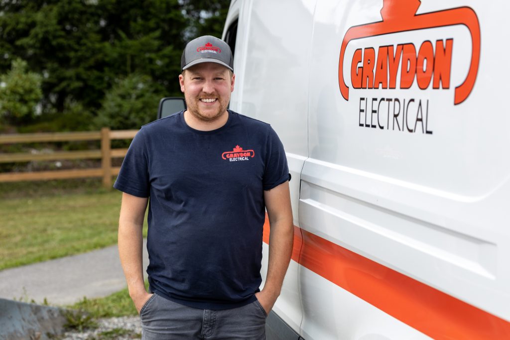 Spruce Tree Media A man in a Graydon Electrical shirt and cap stands smiling beside a company van labeled Graydon Electrical, captured in a grassy outdoor area with a wooden fence in the background. This photograph perfectly frames the essence of professional charm and service. Web Design Videography, Photography Web Development