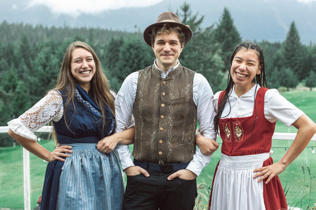 Spruce Tree Media Three people smiling and standing outdoors amidst a picturesque backdrop of trees and mountains capture the essence of Bavarian tradition. Two women in dirndls and a man in a vest and hat pose on a balcony for memorable photography, their arms wrapped around each other. Web Design Videography, Photography Web Development