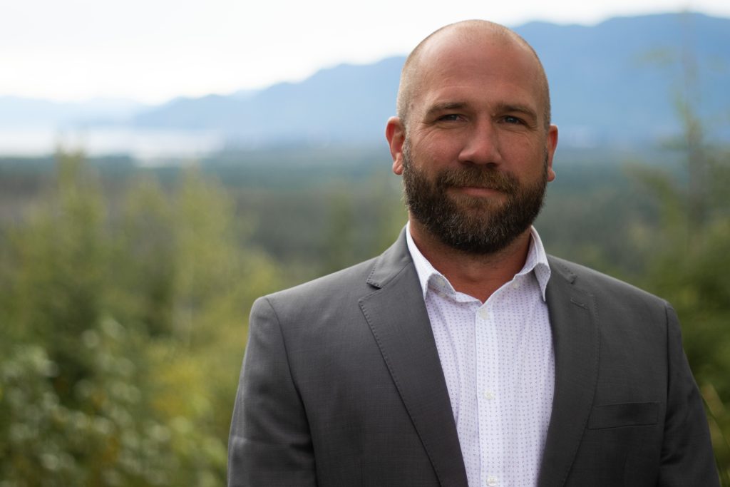 Spruce Tree Media A man with a beard stands outdoors in a gray suit and white shirt, smiling slightly. Captured through skilled photography, the background features a blurred landscape with green trees and mountains under a cloudy sky. Web Design Videography, Photography Web Development